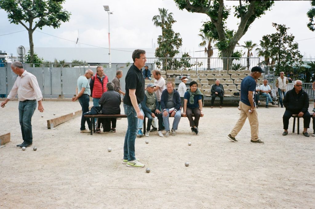 A diverse group of men playing petanque on an outdoor sports ground in a city environment.